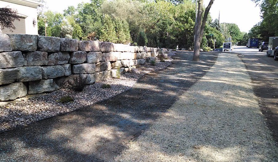 Stone wall alongside a gravel pathway, leading toward trees and a building.