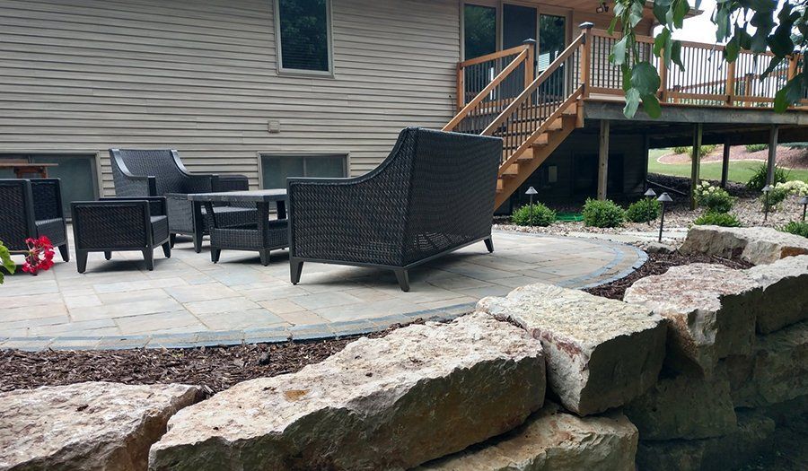 Patio with wicker furniture, stone retaining wall, and house with a wooden deck and staircase.