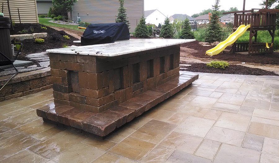 Outdoor brick grilling station on a stone patio, covered grill, and a children's playset in the background.