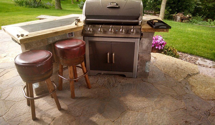 Outdoor grilling station with two stools on a stone patio. A gas grill is centered with a counter on the left.