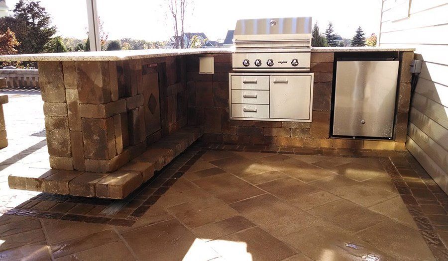 Outdoor kitchen with stainless steel grill, cabinets, and refrigerator built into a brick structure on a patio.