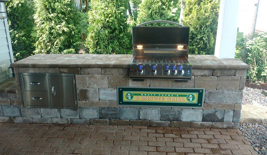 Outdoor kitchen with built-in grill, stone counter, stainless steel storage, and green sign on brick patio.