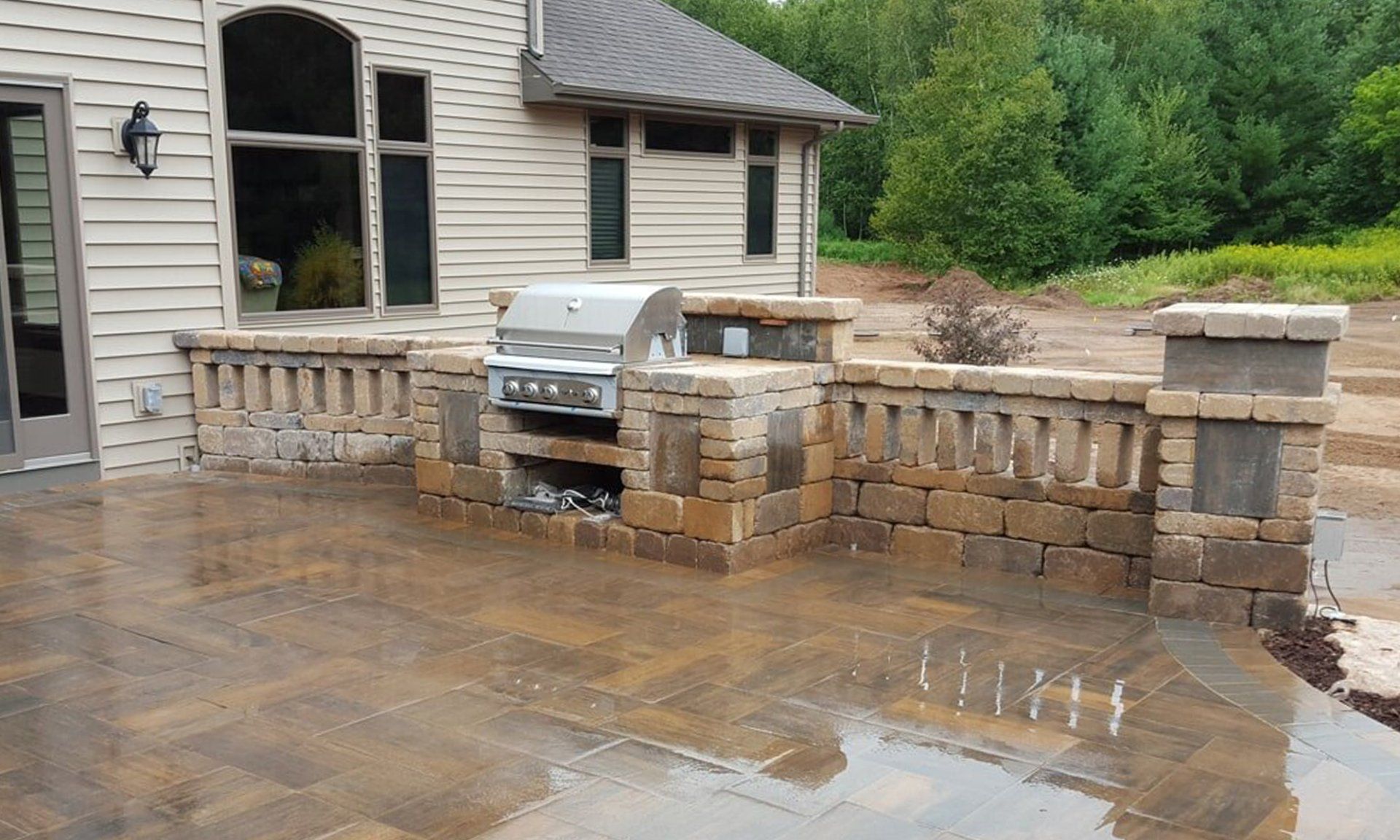 Outdoor kitchen with built-in grill and brickwork, on a paved patio beside a house.