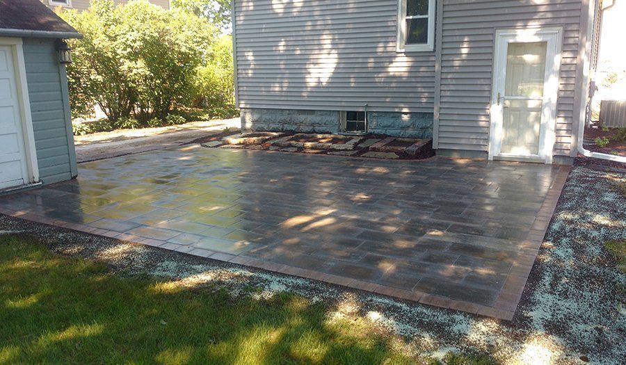 Brick patio next to a light blue house and garage, with grass and gravel borders.
