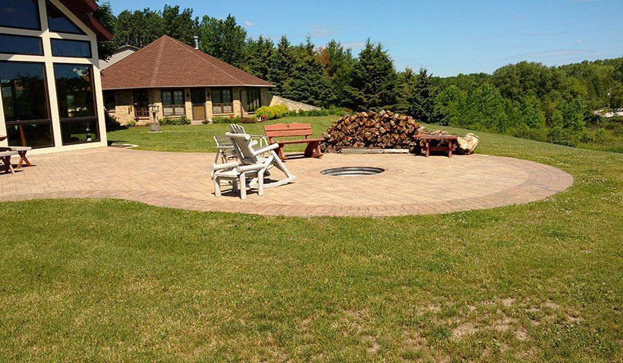 Circular stone patio with fire pit, chairs, and woodpile, next to a house and green lawn under a sunny sky.