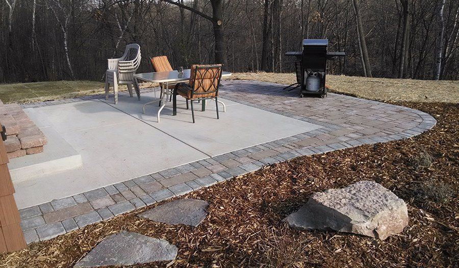 Patio with table, chairs, and grill; bordered by pavers and mulch. Two large rocks in foreground.