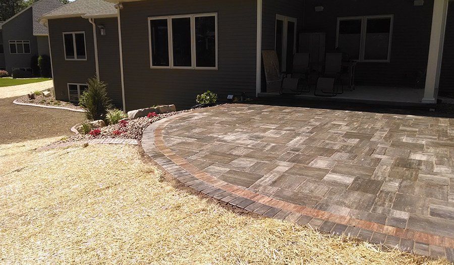 A brick patio with a circular edge next to a house with dark gray siding. Brown mulch and dry grass surround it.