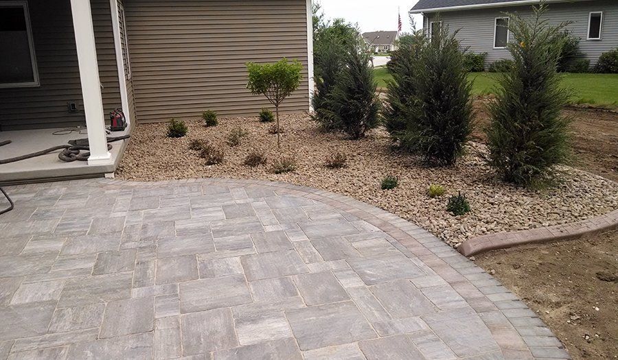 Paver patio next to a gravel bed with small trees and bushes; home in the background.