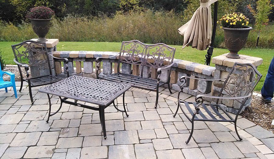 Patio with wrought iron furniture: table, chairs, bench on stone pavers near a brick wall, urns, and umbrella.