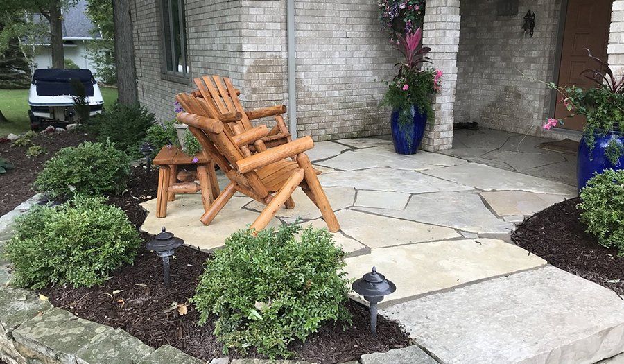 Wooden chairs on a stone patio surrounded by greenery and mulch in front of a brick building.