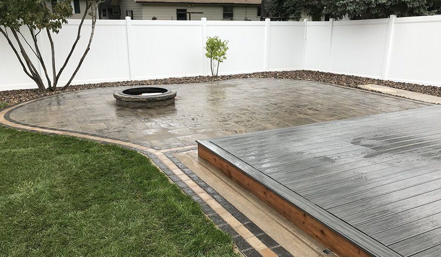 Rain-covered patio with brick pavers, fire pit, wooden deck, and white fence.