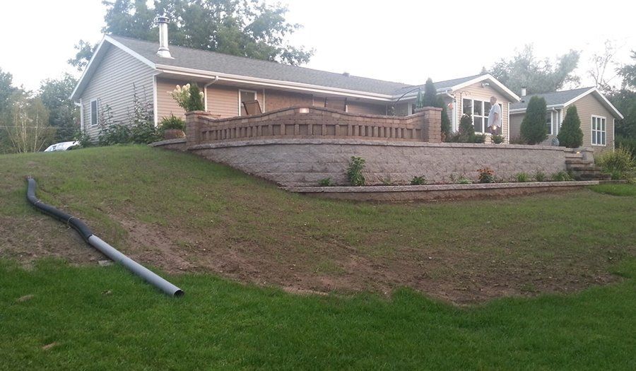 A house with a brick retaining wall on a grassy hill; a drainage pipe is in the foreground.