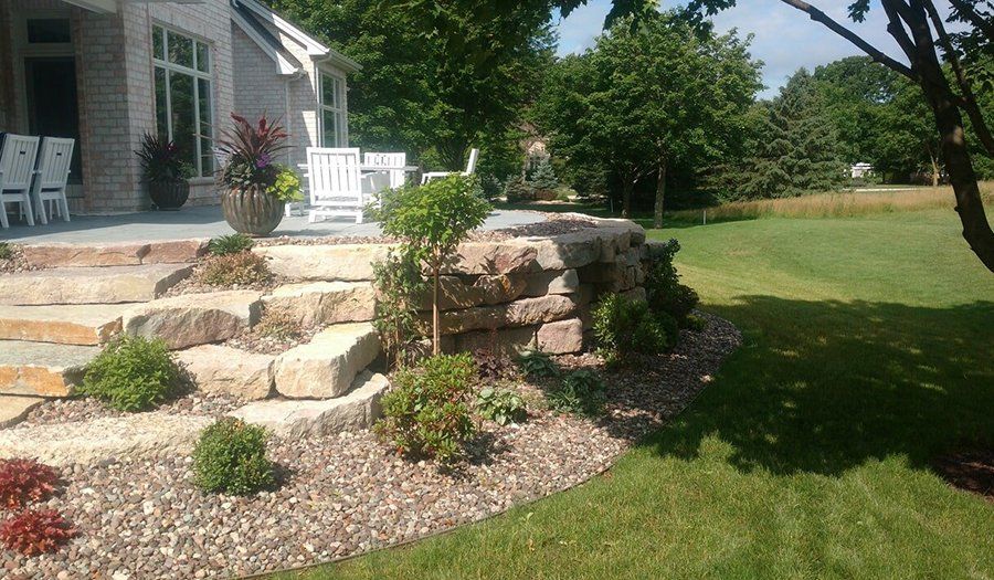 Stone steps and retaining wall leading to a patio with outdoor furniture, surrounded by landscaping and a grassy lawn.