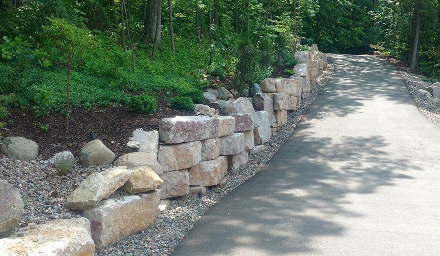Stone retaining wall along a paved path in a wooded area.