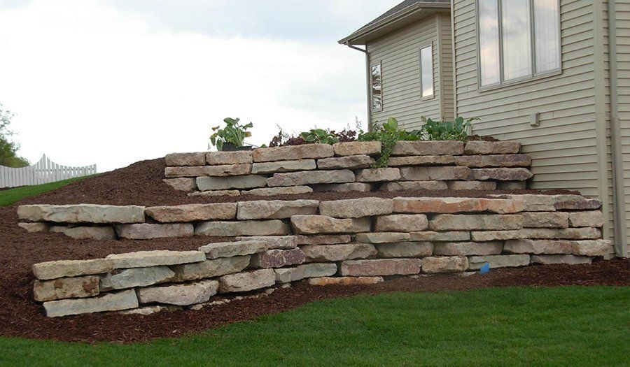 Stone retaining wall with plants near a house, on a grassy lawn. Brown mulch.