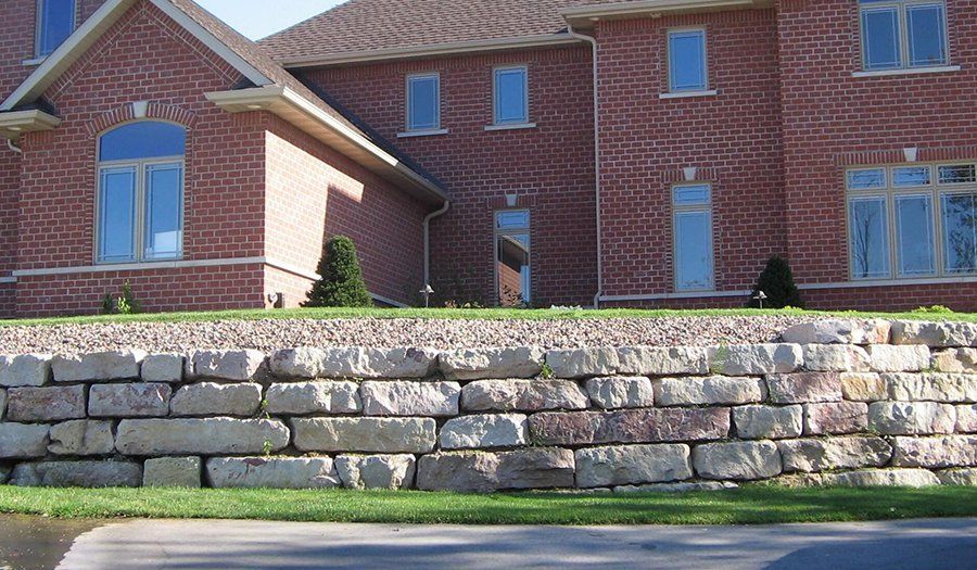 Stone retaining wall in front of a brick house with grass and small trees.