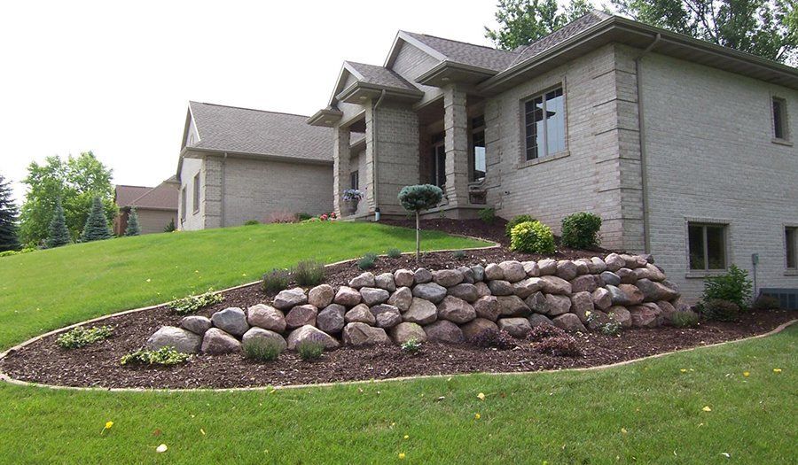 Stone retaining wall with plants in front of a light brick house on a grassy hill.