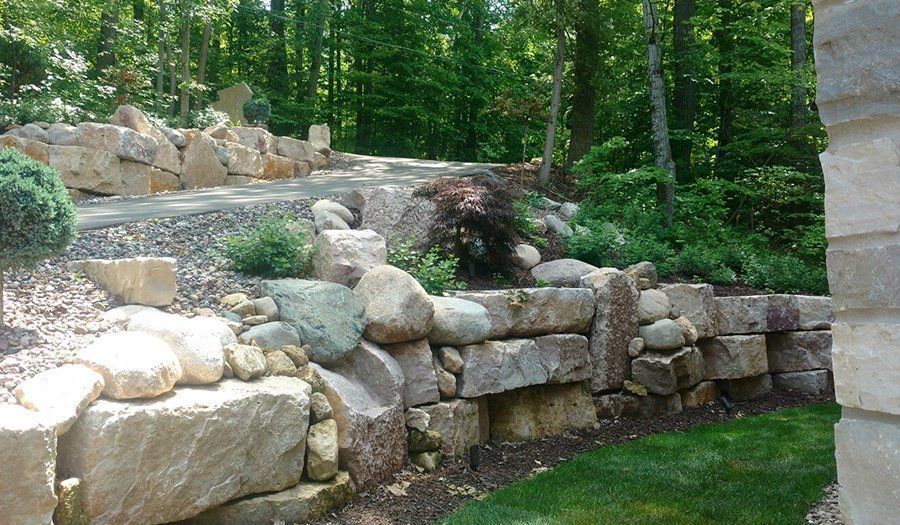 Stone retaining wall with pathway, lush greenery, and trees in background.