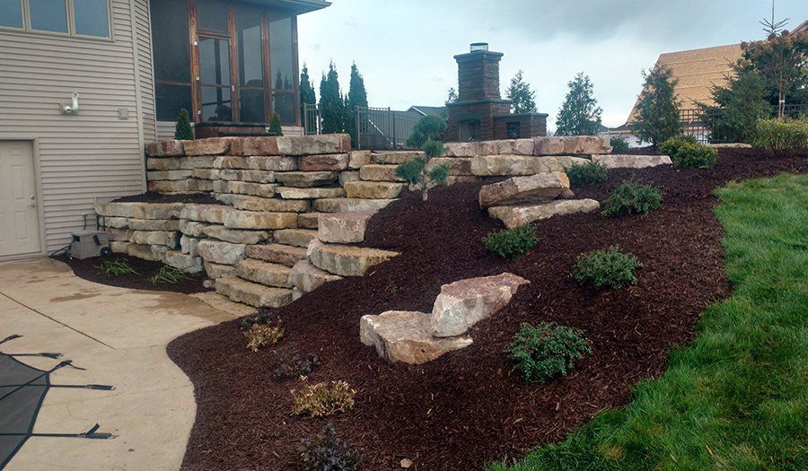 Stone retaining wall steps with brown mulch and green shrubbery leading to a house.