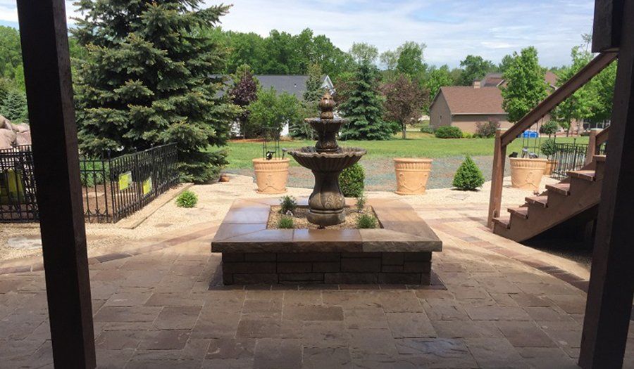 Fountain on a stone patio with potted plants, surrounded by a lawn and houses in the background.