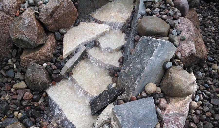 Water flows over irregularly-shaped, light-colored stones surrounded by pebbles and larger rocks of various colors.