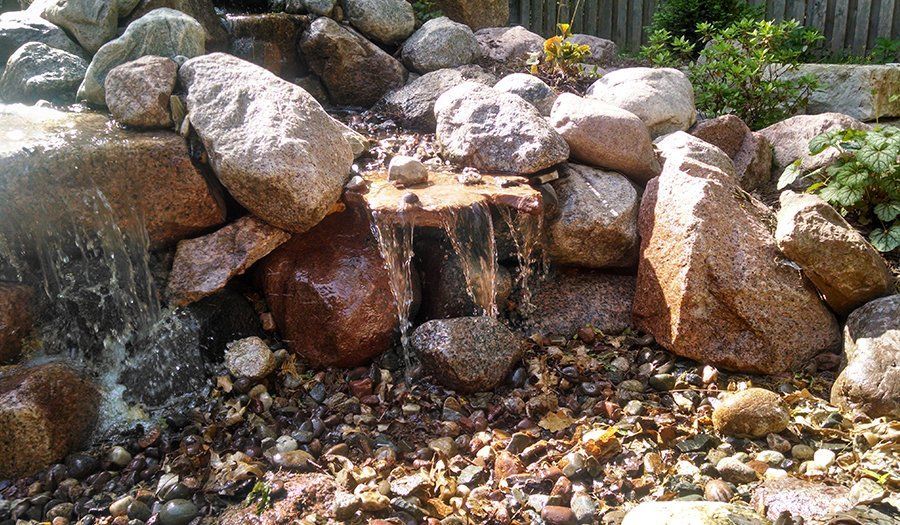 Small waterfall cascading over rocks in a garden setting, with sunlight reflecting on wet surfaces.