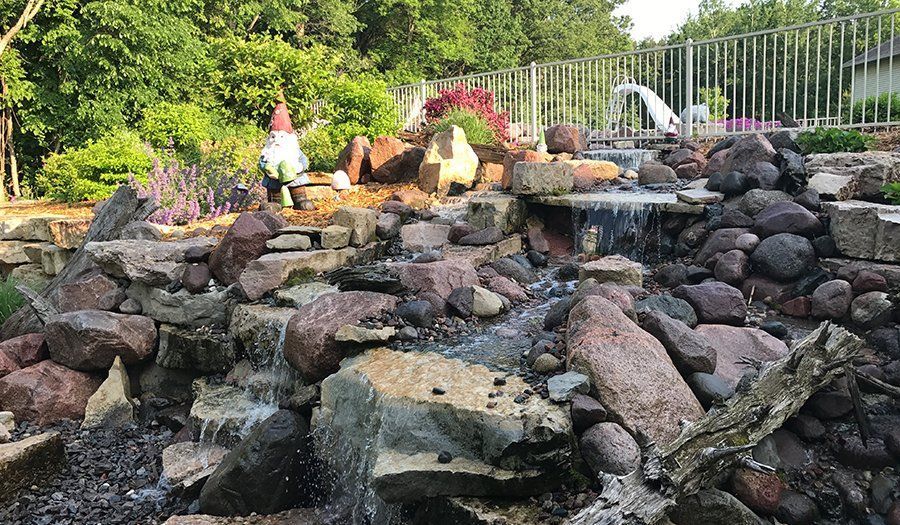 Water cascading down rocks in a garden. A gnome sits near flowers; a pool and fence are in the background.