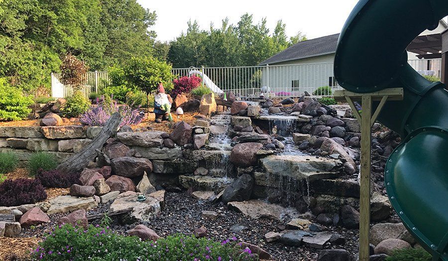 A rocky waterfall feature with flowing water, plants, and garden gnomes. A green slide is in the foreground.