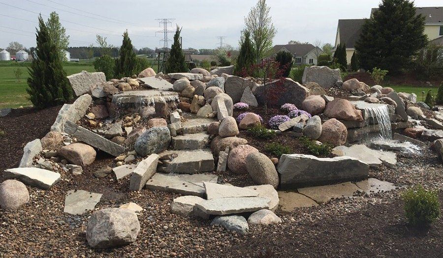 Rock waterfall feature in a yard, with layered stones, small trees, and surrounding brown mulch.