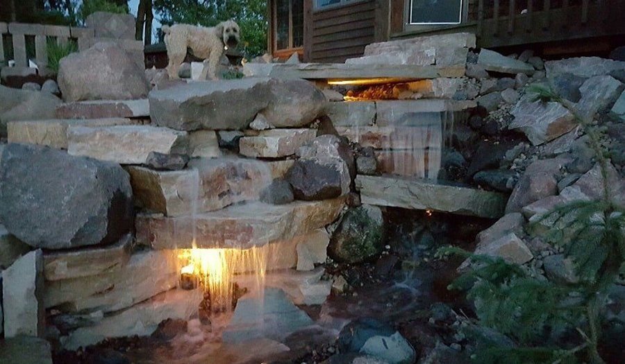 A multi-tiered rock waterfall with water flowing, illuminated by lights, and a dog standing at the top.