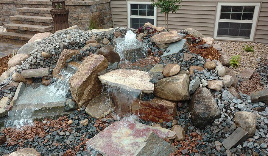 Water cascading down a rocky waterfall feature in a garden, near a house with windows.