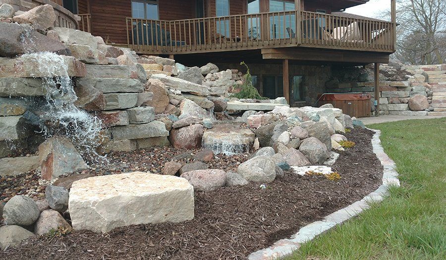 Waterfall feature in front of a log cabin with a deck, surrounded by rocks and landscaping.