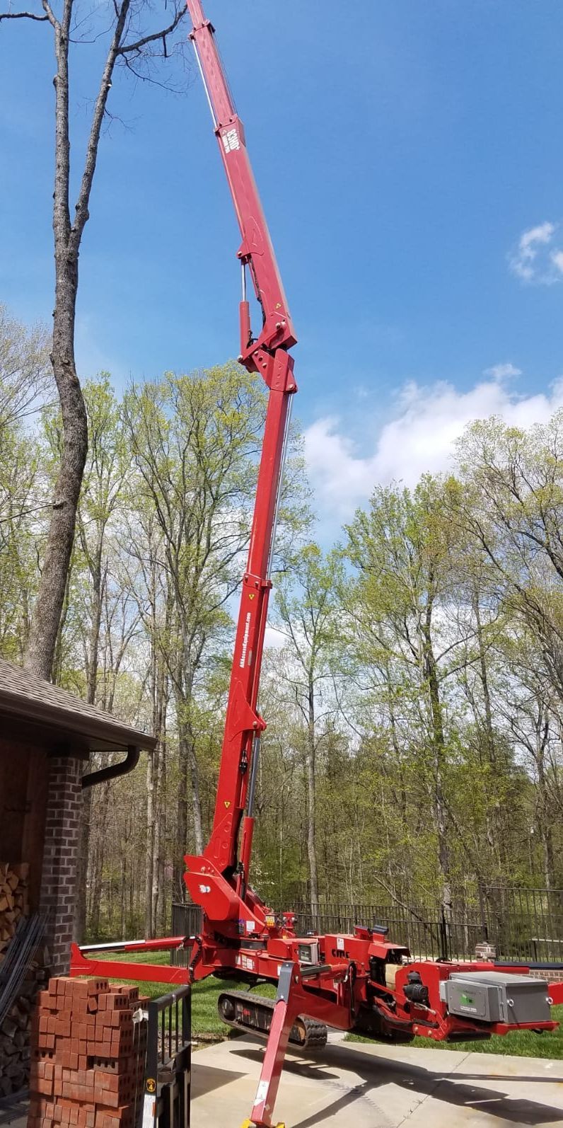 Red crane boom extended over a brick building in a wooded yard under a blue sky.