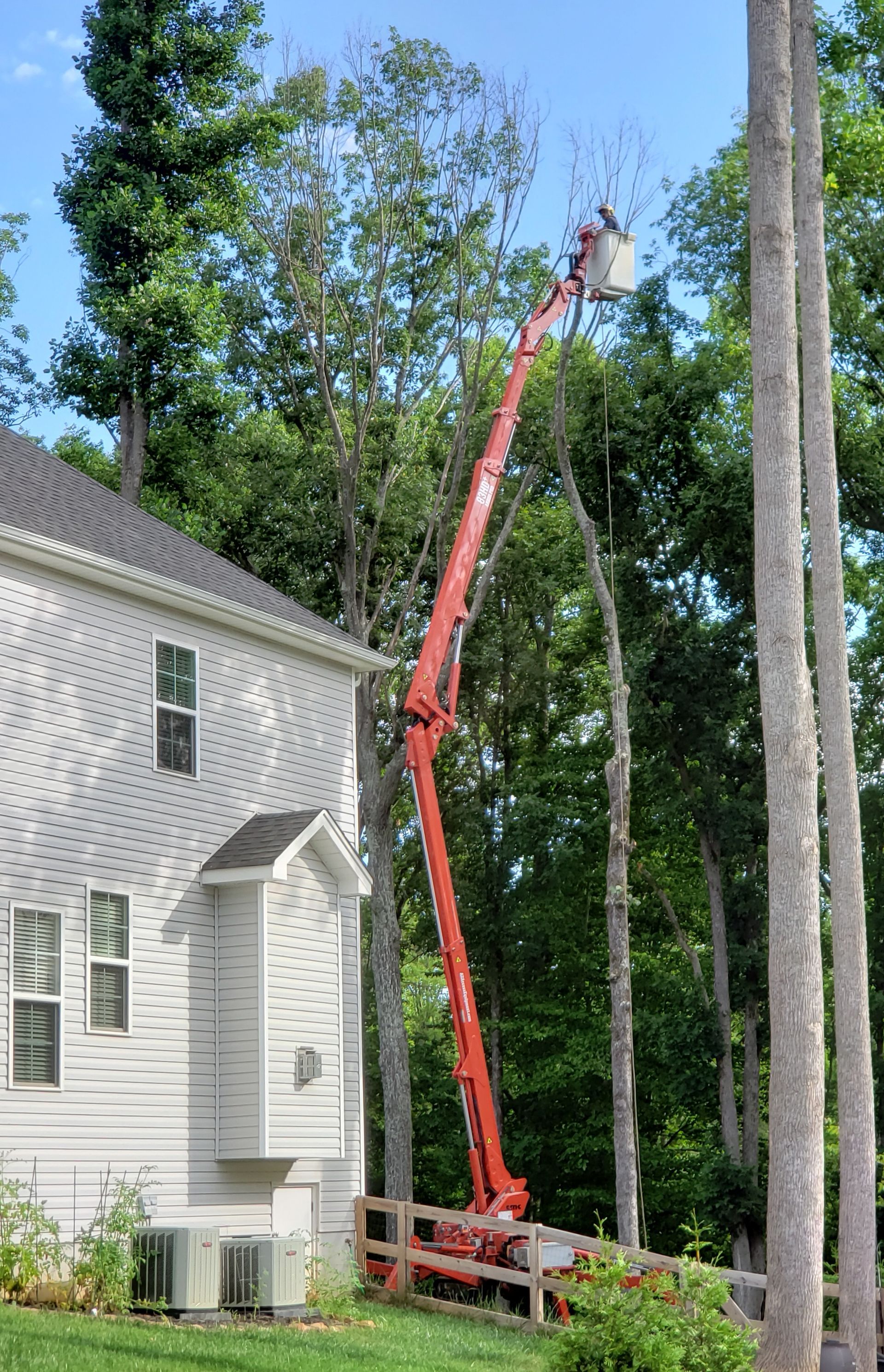 Red boom lift extended beside a white house, reaching up toward tall trees.