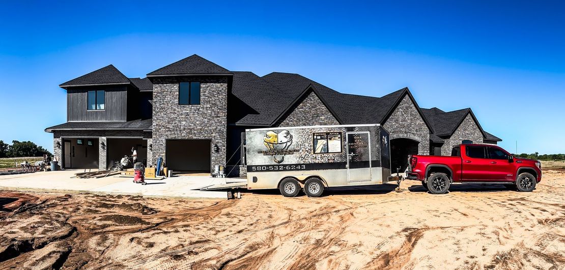 New house under construction, red pickup truck towing a trailer on a dirt lot, clear blue sky.