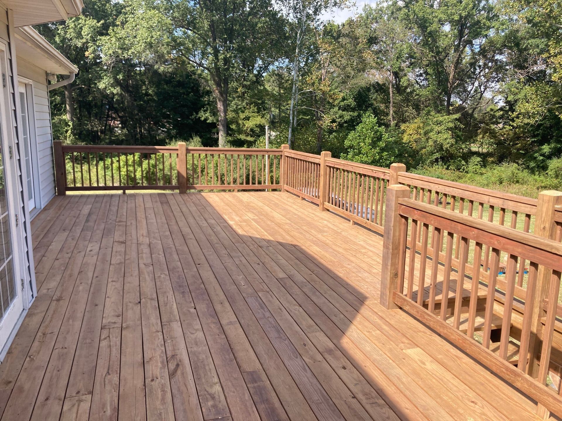 Wooden deck with railings, connected to a white house with a wooded background.