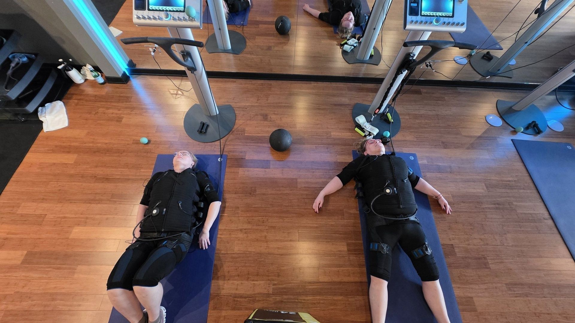 Two people lying on mats in a gym, doing exercise with weighted machines around them.