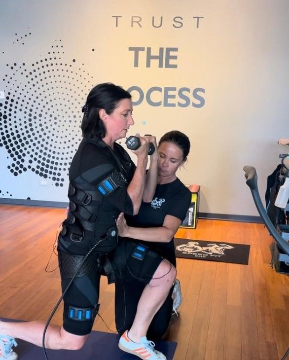 Two women in workout gear practice a training hold in a studio with “TRUST THE PROCESS” on the wall.
