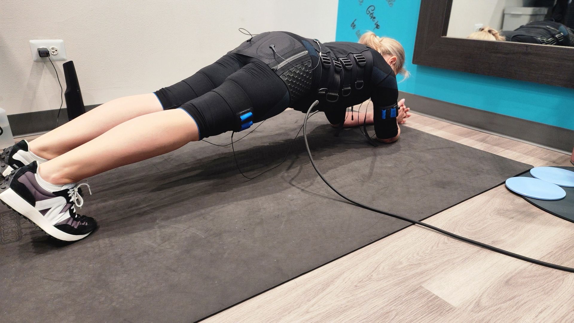Person doing a plank exercise on a black mat in a gym, wearing black workout clothes and shoes.