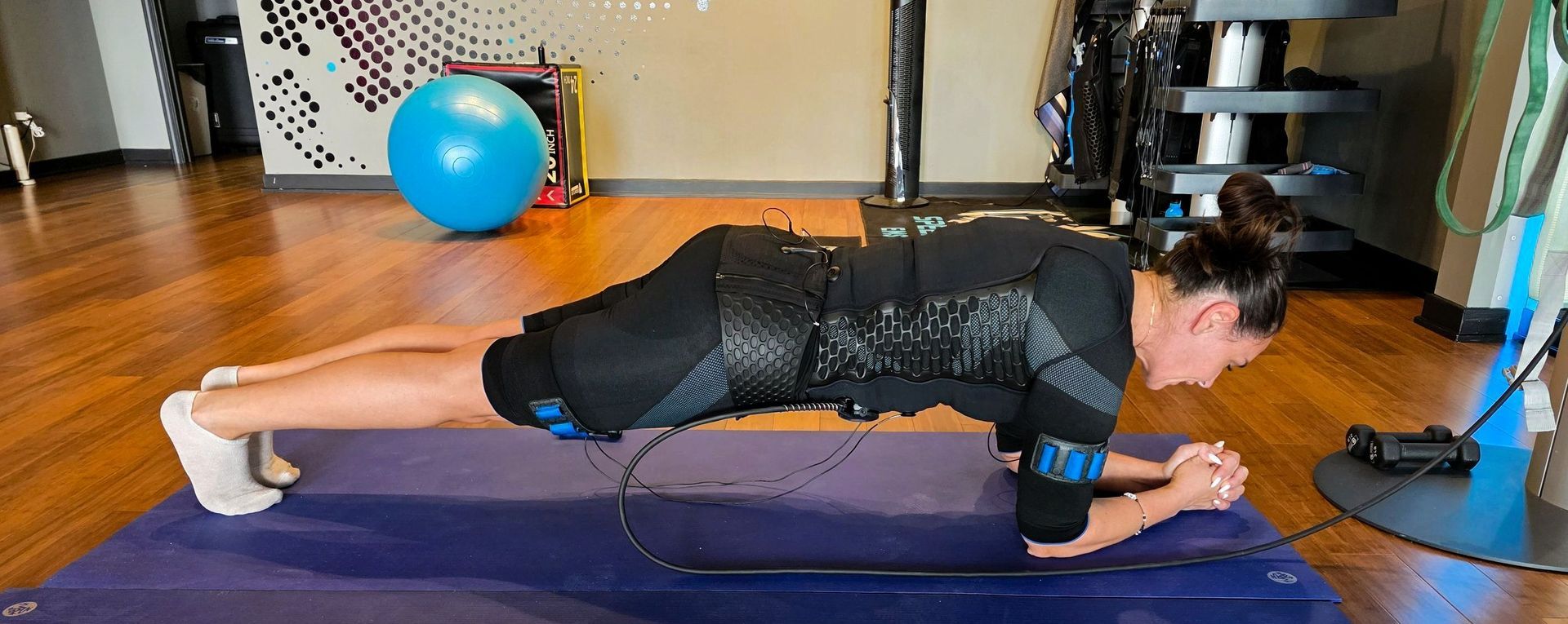 Person holding a forearm plank on a mat in a home gym, with a blue exercise ball in the background