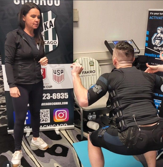 Two people at a gym, one squatting while holding a weighted bar facing a woman standing nearby.