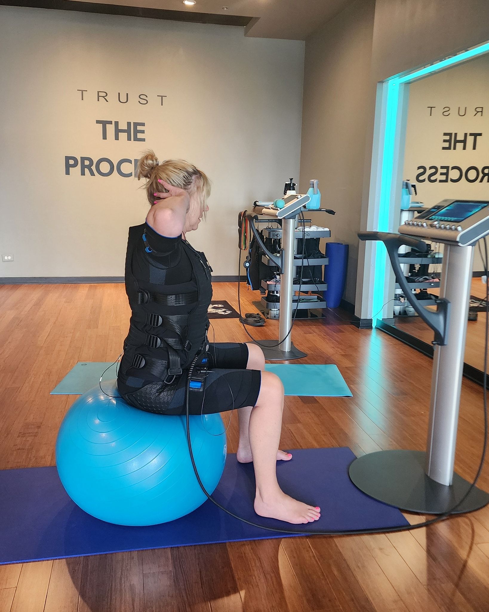 Person seated on a blue stability ball in a gym, beside workout equipment and turquoise yoga mats.