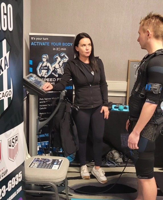 Woman in black athletic wear talks to a trainer in an EMS workout studio.