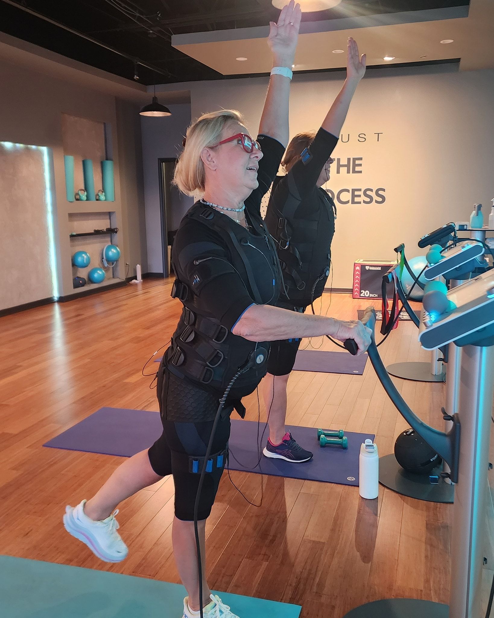 People exercising in a gym, lifting one leg and reaching up beside workout machines.