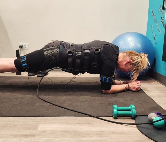 Person in black EMS suit doing a forearm plank on a mat in a gym, with exercise balls and dumbbells nearby