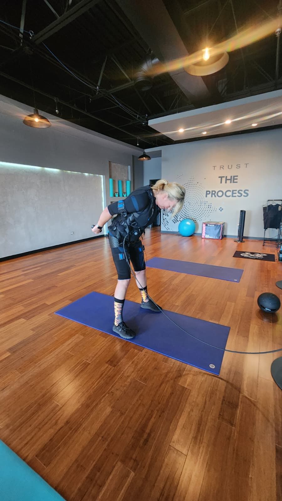 Person exercising on a blue mat in a gym studio with wood floors and exercise equipment in the background
