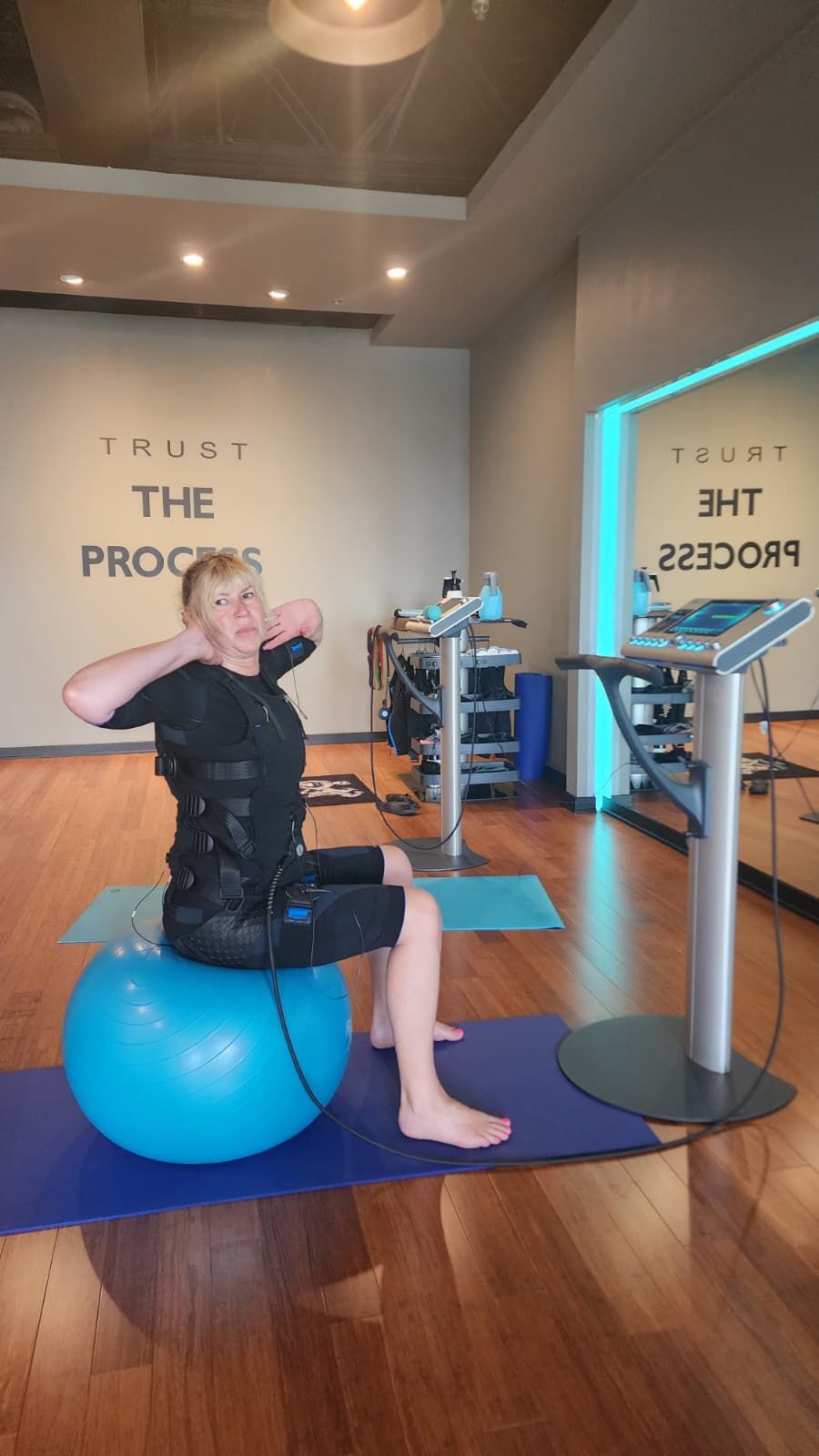 Person seated on a blue exercise ball in a gym, stretching an arm beside cardio machines.