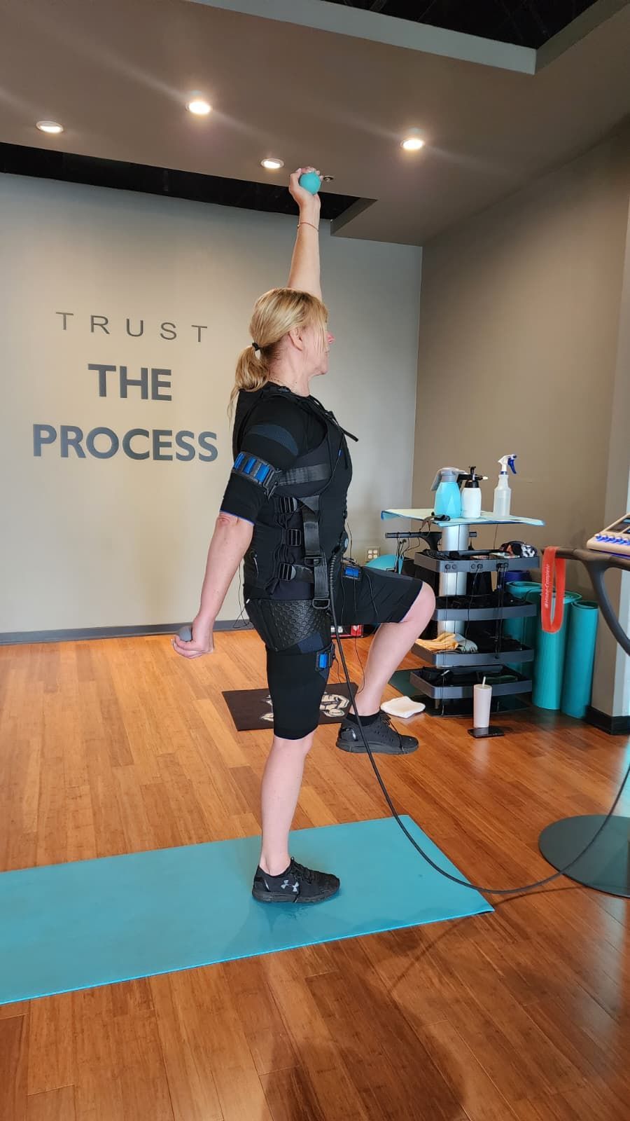Woman holding a dumbbell overhead on a blue mat in a gym, with “TRUST THE PROCESS” on the wall.