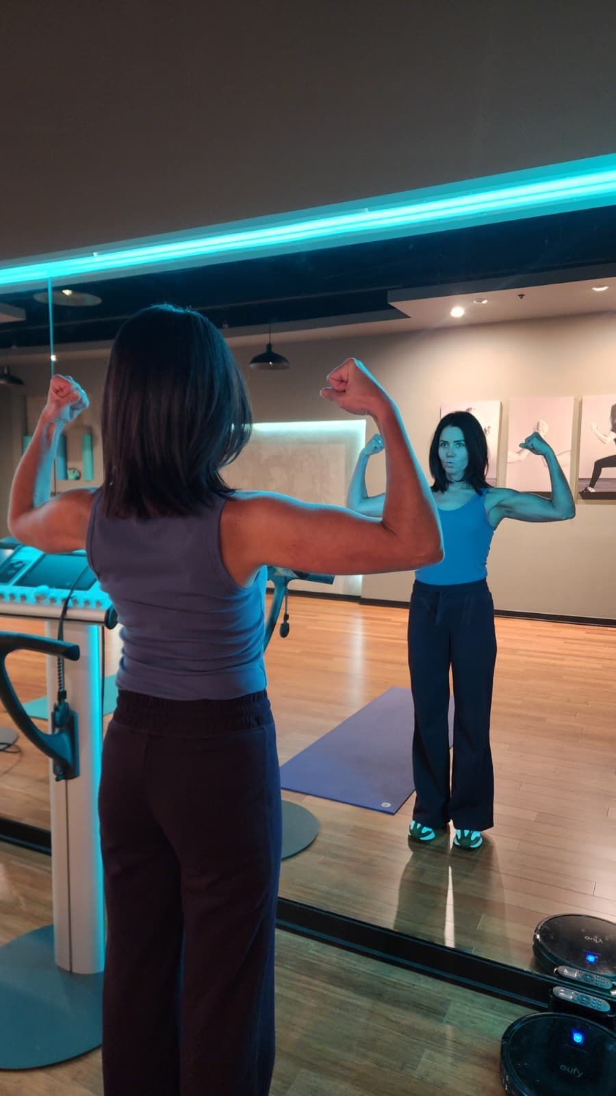 Person in a mirrored studio raises flexed arms during a workout, with blue lighting and a yoga mat.