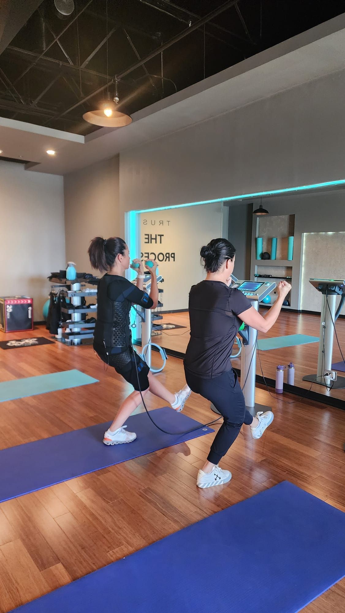Two people doing a workout in a mirrored studio on blue mats, one holding weights.